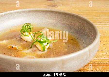 Gnocchi wonton in brodo dorato saporito, guarniti con nastri di scalogno arricciati, fiocchi di peperoncino e condimento a base di panna in una ciotola di ceramica. Foto Stock