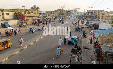 Vista aerea di una scena stradale affollata a Kano con persone che camminano, motociclette e auto che percorrono la strada polverosa fiancheggiata da negozi e edifici locali, Foto Stock