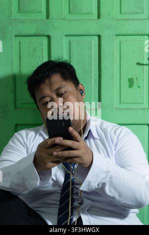 Un giovane uomo asiatico sovrappeso concentrato digita su uno smartphone nero mentre è seduto contro una porta di legno verde rustica. Il professionista aziendale remoto w Foto Stock