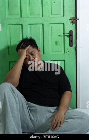 Un uomo asiatico adulto è seduto su un letto bianco mentre appoggia pesantemente una mano sulla fronte, esprimendo un forte senso di stress, mal di testa o affaticamento mentale. Foto Stock