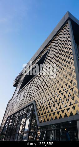 Edificio moderno che brilla sotto un cielo blu terso. Motivi geometrici coprono la sua facciata ruvida. Porte di vetro scuro invitano i visitatori ad entrare Foto Stock