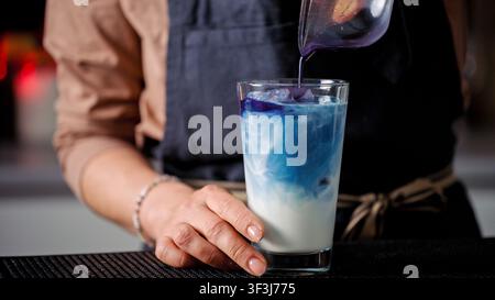 Versare tè ai fiori di farfalla viola nel latte freddo a un bancone da caffè professionale durante il giorno Foto Stock