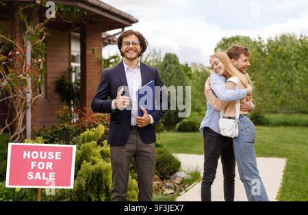 Agente immobiliare felice mostrando pollice in su gesto mentre giovane coppia abbracciando vicino alla loro nuova casa, fuori Foto Stock
