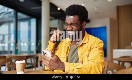 Uomo afroamericano felice che celebra il successo guardando il smartphone in un caffè. Uomo che alza il pugno e ride con entusiasmo mentre controlla buone notizie Foto Stock