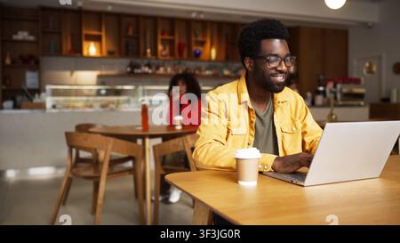 Uomo afroamericano concentrato con occhiali che lavora su un laptop in un caffè e beve una bevanda calda. Uomo seduto a un tavolo di legno con una tazza di caffè, donna Foto Stock