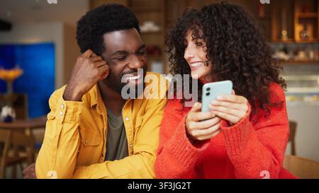 Un uomo e una donna condividono un momento gioioso, sorridendo e guardando lo schermo di un telefono cellulare in un accogliente ambiente da caffè Foto Stock