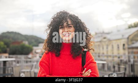 Ritratto di donna caucasica sorridente con capelli ricci in piedi all'aperto vicino ad edifici e sorridente. Donna in maglione luminoso che guarda la telecamera con Foto Stock