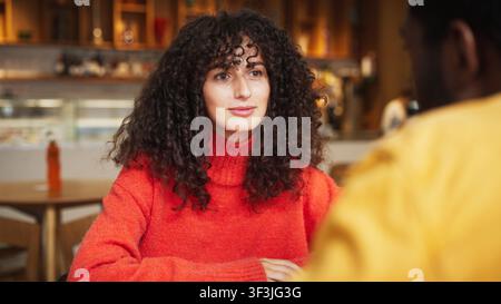 Donna caucasica con capelli ricci appoggiata su una mano mentre parla al chiuso di un caffè. Donna impegnata in una piacevole conversazione con un uomo afroamericano Foto Stock
