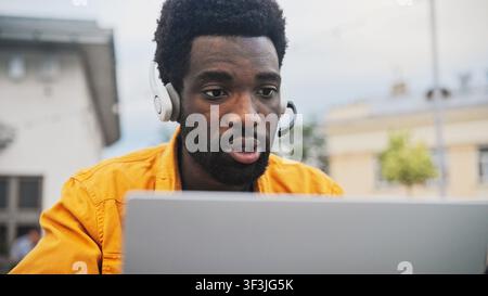 Affascinante uomo afroamericano con barba che indossa un auricolare e parla mentre lavora su un laptop all'aperto. Uomo che comunica online, seduto vicino a un ambiente moderno Foto Stock