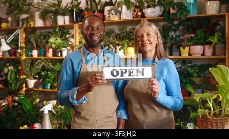 Ritratto di due fioristi di successo e allegri che guardano l'obiettivo sorridendo, tenendo un cartello "Aperto", indossando un grembiule e godendosi il lavoro al negozio di fiori. Giovane africano Foto Stock