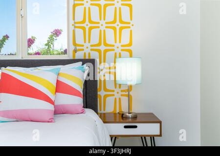 Una scena allegra della camera da letto. Un letto adornato con cuscini a righe colorati si trova accanto a un comodino in legno e a una vista su una vegetazione lussureggiante dalla finestra. L'int della stanza è int Foto Stock