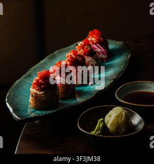 Vista laterale foto professionale di rotoli di sushi gourmet guarniti con caviale tobiko rosso su un piatto turchese, con salsa di soia e wasabi. Foto Stock