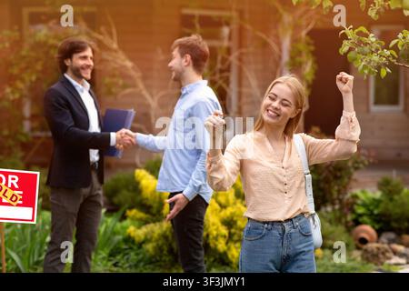 Felice giovane donna con la chiave di casa e suo marito scuotendo mani con il realtor in cortile Foto Stock