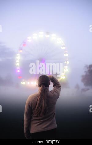 Vista di una donna in piedi nella fitta nebbia che guarda una ruota panoramica luminosa di notte a Berna, Canton Berna, Svizzera. Foto Stock
