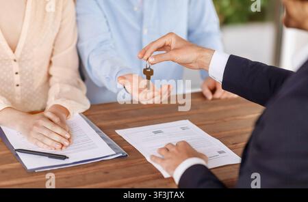 Agente immobiliare che dà la chiave della casa a giovane coppia dopo aver firmato l'accordo di proprietà al tavolo, primo piano Foto Stock