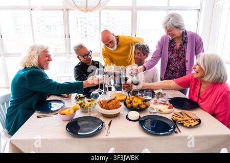 Gruppo vario di amici anziani sorridenti che godono di una cena gioiosa e brindano con vino rosso, celebrando l'amicizia e la solidarietà in una luce Foto Stock