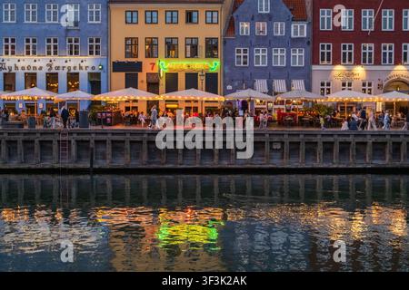 Vista degli edifici storici colorati e dei ristoranti lungo il lungofiume Nyhavn a Copenaghen, Danimarca, con persone che cenano ai tavoli e una serata calda Foto Stock