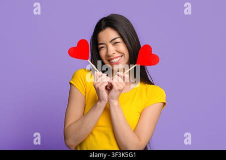 Giocosa donna asiatica in posa con due cuori di carta in mani, sfondo viola Foto Stock