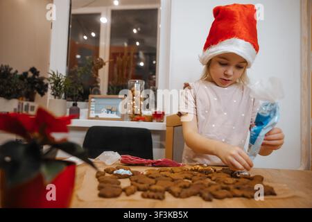 Bambino decora biscotti di zenzero durante la stagione delle feste con un cappello festivo in una cucina accogliente Foto Stock