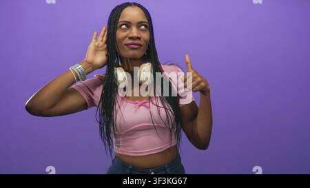 Donna con cuffie intorno al collo che indossa un crop top rosa, tiene la mano all'orecchio e lancia un'occhiata di lato in uno studio viola; approvazione curiosa. Foto Stock