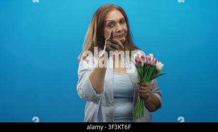 Una donna anziana che tiene in mano tulipani rosa tocca il tempio e punta il dito in studio; riflesso premuroso. Foto Stock