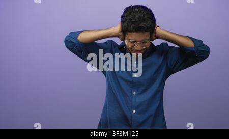Giovane ispanico che indossa occhiali e camicia blu che preme entrambe le mani alle orecchie in uno studio; stress disagio. Foto Stock