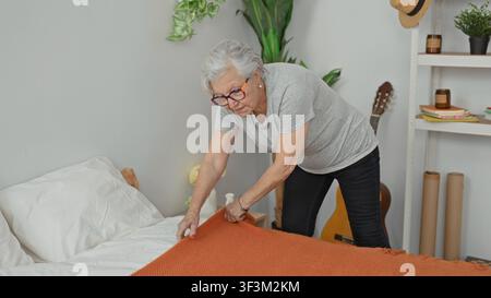 Anziana che liscia una coperta arancione sul letto con le mani mentre si china nella camera da letto; indipendenza calma. Foto Stock