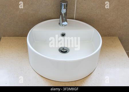 Un lavandino da bagno contemporaneo e pulito con un bacino bianco rotondo, un rubinetto cromo elegante e un piano di lavoro beige strutturato Foto Stock