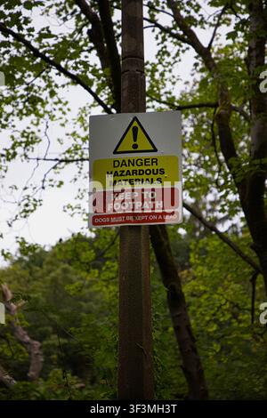 Un chiaro cartello di sicurezza montato su un palo che recita Pericolo Materiali Pericolosi - Rimanere sul marciapiede - I cani devono essere tenuti al guinzaglio. Foto Stock