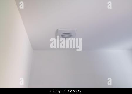Un ventilatore di scarico bianco è montato su un soffitto bianco e liscio, fornendo ventilazione a una stanza Foto Stock