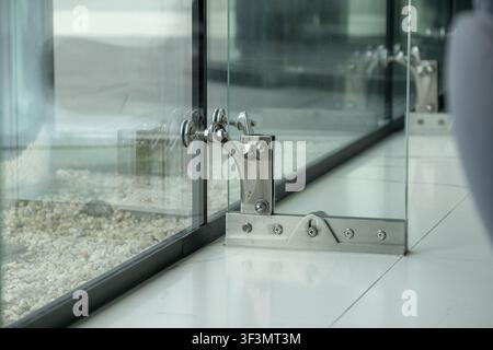 Primo piano di ferramenta in acciaio inossidabile che fissa una porta in vetro senza telaio in un edificio contemporaneo. Dettaglio architettonico moderno con G montato a pavimento Foto Stock