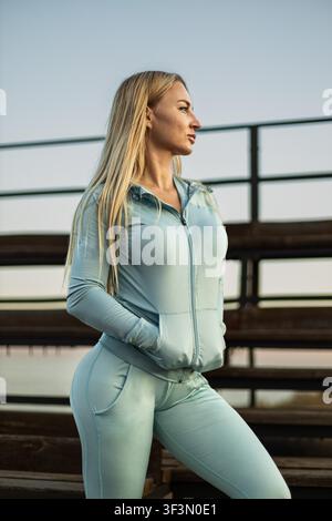Ritratto di profilo di una giovane donna caucasica atletica con lunghi capelli biondi che indossa una tuta sportiva azzurra, in piedi all'aperto sulle tribune di legno dello stadio durante il tramonto. Foto Stock