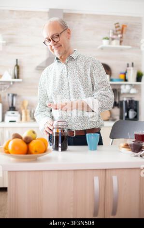 Un uomo anziano prepara il caffè con una French press durante la colazione in cucina, godendosi una bevanda mattutina rilassata con una tazza vintage, con la preparazione casalinga, la routine della colazione e il tempo libero rilassato degli anziani. Foto Stock