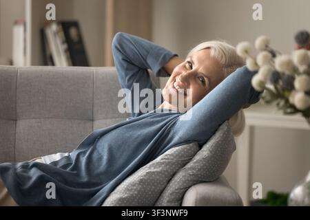 Donna anziana serena sdraiata sul divano con le mani dietro la testa Foto Stock