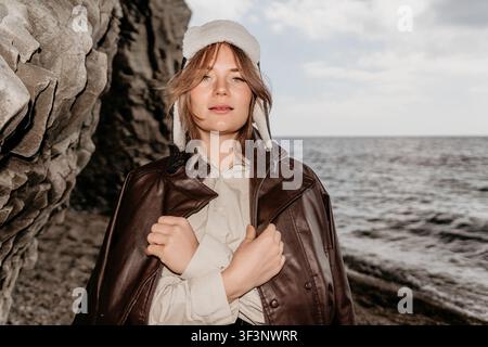 Ritratto su roccia oceanica di una giovane donna con giacca di pelle e cappello invernale sulle scogliere del mare in una giornata nuvolosa Foto Stock