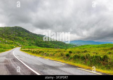 Strada asfaltata rurale curva sul Wild Atlantic Way in Irlanda che attraversa colline verdi lussureggianti e pendii rocciosi sotto un cielo nuvoloso pesante. La vuota Foto Stock