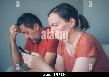 Una coppia di mezza età seduta fianco a fianco, la donna assorbita dal suo smartphone, illustrando distanza emotiva e distrazione digitale. L'immagine trasmette il tema Foto Stock