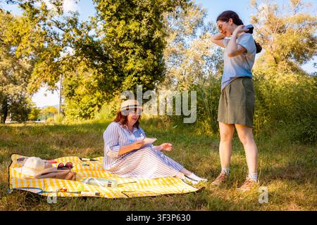 Madre e figlia adulta trascorrono del tempo insieme durante un picnic estivo nel parco, godendosi snack, sole e interazioni familiari giocose nella natura. Largo Foto Stock