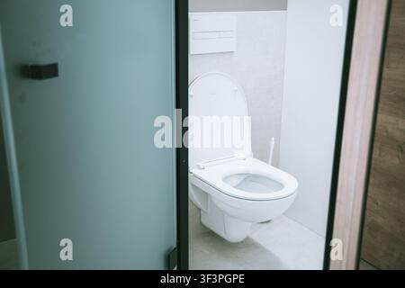 Un interno di bagno pulito e moderno con una toilette bianca a muro con coperchio aperto, incorniciata da una porta in vetro smerigliato accanto a una luce minimalista Foto Stock