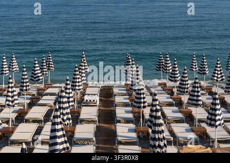 Vista geometrica ad angolo alto di file di ombrelloni blu a righe chiusi e lettini su una spiaggia di ciottoli vuota vicino al mare nella Costa Azzurra francese. Foto Stock