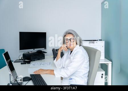 Medica anziana con camice da laboratorio e occhiali, seduta alla sua scrivania Foto Stock