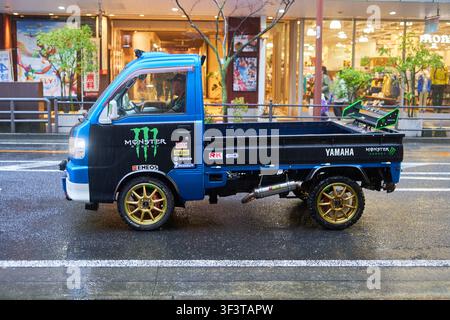 Un mini camioncino Kkei giapponese modificato blu e nero parcheggiato su una strada urbana bagnata davanti ad edifici commerciali. Il veicolo è dotato di una lega dorata Foto Stock