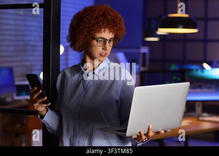 Giovane donna d'affari con capelli rossi ricci e occhiali che lavora fino a tardi in un ufficio moderno, tiene un laptop e uno smartphone, Tiene un laptop in una mano a Foto Stock