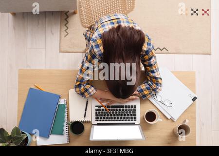Giovane donna oberata di lavoro che dorme sulla tastiera di un laptop alla scrivania dell'ufficio domestico, concetto di burnout e affaticamento, vista dall'alto Foto Stock