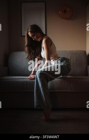Giovane donna seduta tranquillamente su un divano in un appartamento accogliente, illuminato dalla dolce luce del tramonto in arrivo dalla finestra. Interno domestico rilassato e intimo con atmosfera calma, abbigliamento casual, Foto Stock