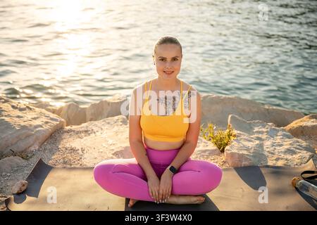 Donna sorridente seduta a gambe incrociate su un tappeto da yoga su scogliera sul mare all'alba, pratica meditazione e respirazione consapevole per sostenere il benessere, la calma e Foto Stock