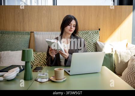 Donna d'affari in un caffè bilancia con maestria lavoro e relax, utilizzando un laptop e godendosi una pausa caffè. La scena cattura l'equilibrio della vita moderna thro Foto Stock