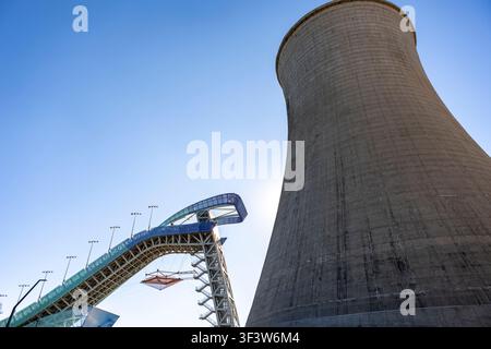 Vista ad angolo basso di una moderna rampa per salti con gli sci accanto a un enorme torre di raffreddamento industriale sotto un cielo blu terso in Cina, evidenziando ingegneria, sport an Foto Stock