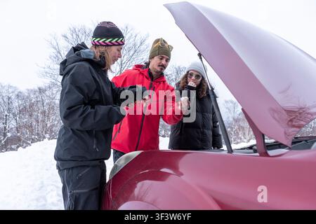 Tre amici riuniti intorno a un'auto con il cofano aperto, ispezionano il motore in un paesaggio innevato. Sono vestiti pesantemente con giacche invernali e cappelli Foto Stock