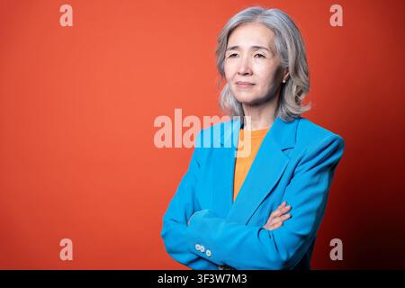 Ritratto in studio di una donna asiatica anziana e sicura di sé con capelli grigi che indossa un blazer blu e un top arancione, con le braccia incrociate, in piedi su uno sfondo arancione brillante Foto Stock